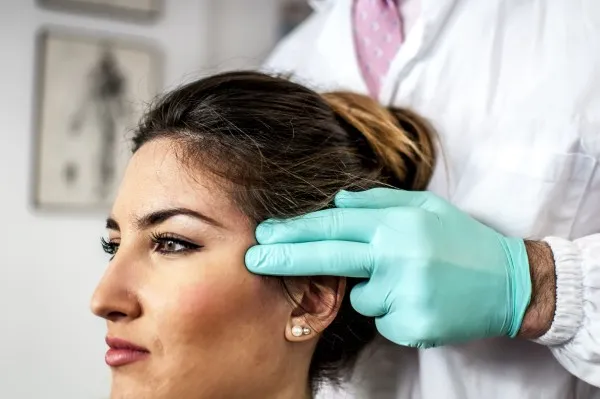 Un doctor, que podría ser un cirujano plástico certificado, realiza un examen táctil en el área de la sien de la paciente.